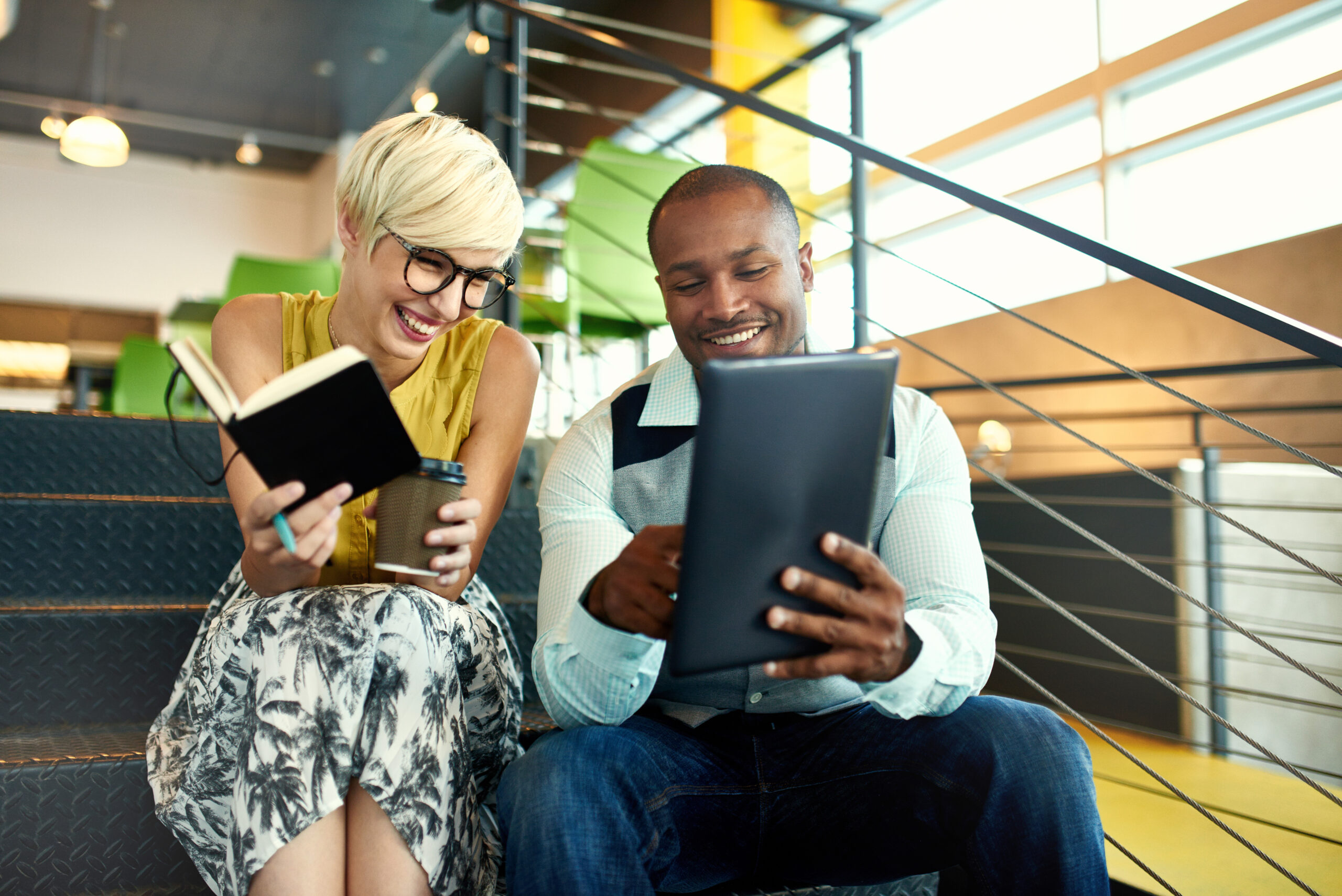 Two creative millenial small business owners working on social media strategy using a digital tablet while sitting in staircase